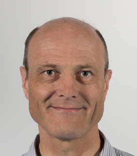 A bald man with a slight smile and wearing a light-colored shirt is looking directly at the camera against a plain background.