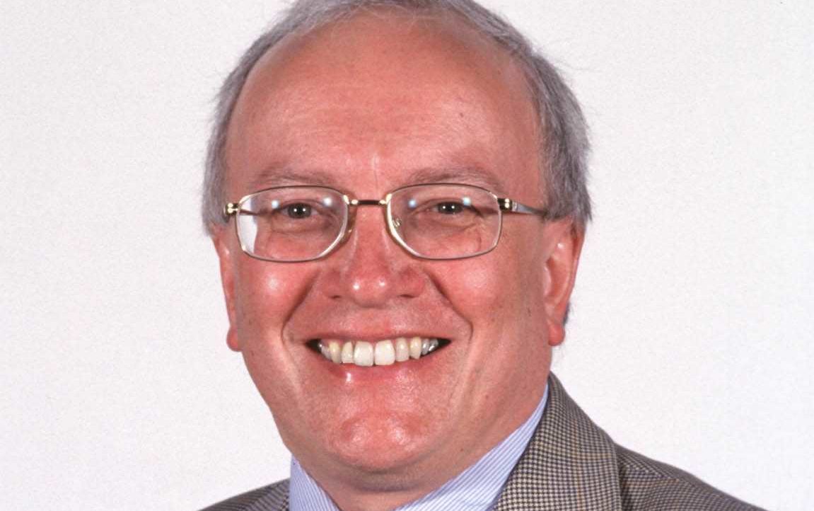 Smiling man with glasses, wearing a checked blazer and a blue shirt, against a plain white background.