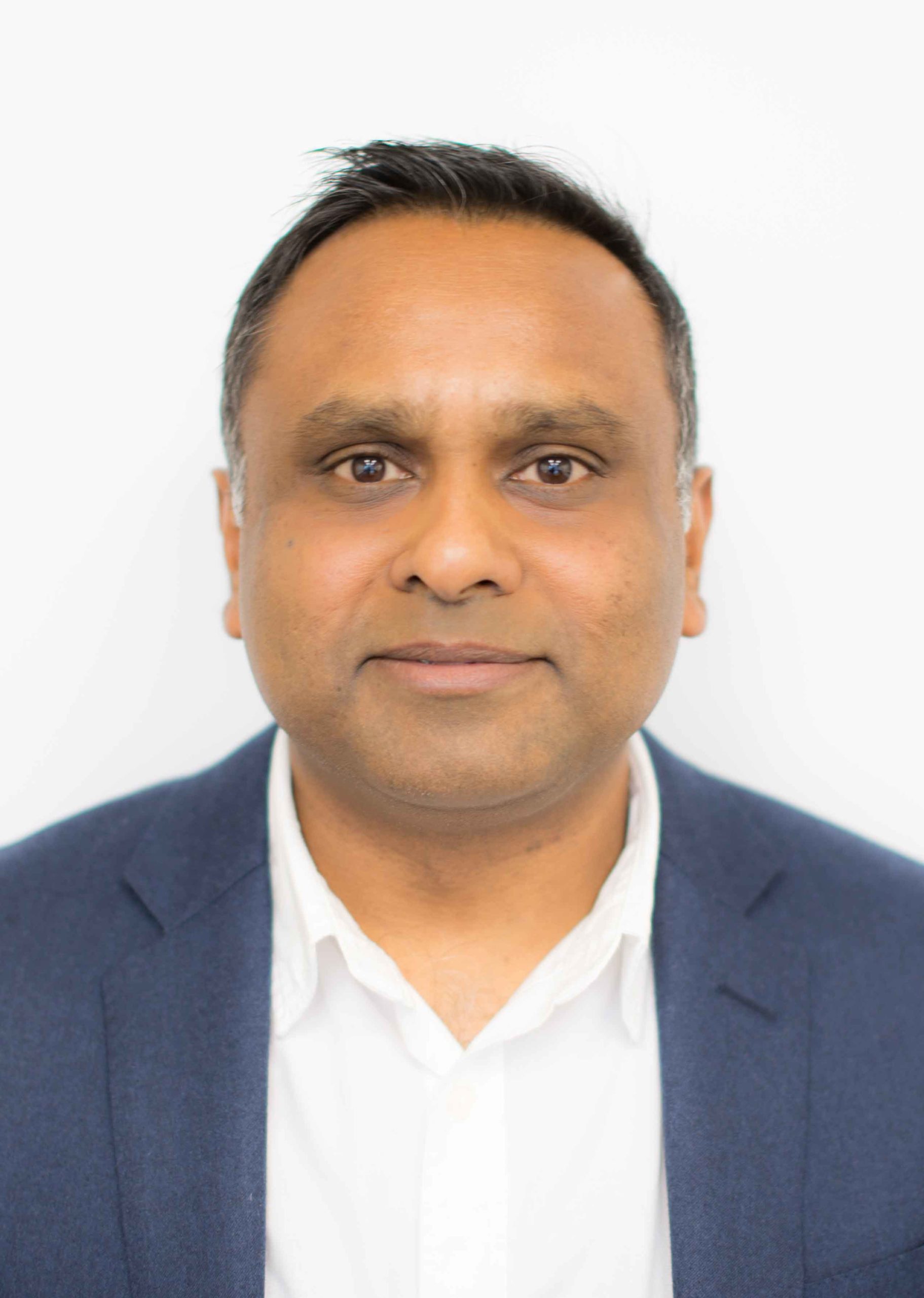Man with short dark hair wearing a white shirt and blue blazer, looking directly at the camera. White background.