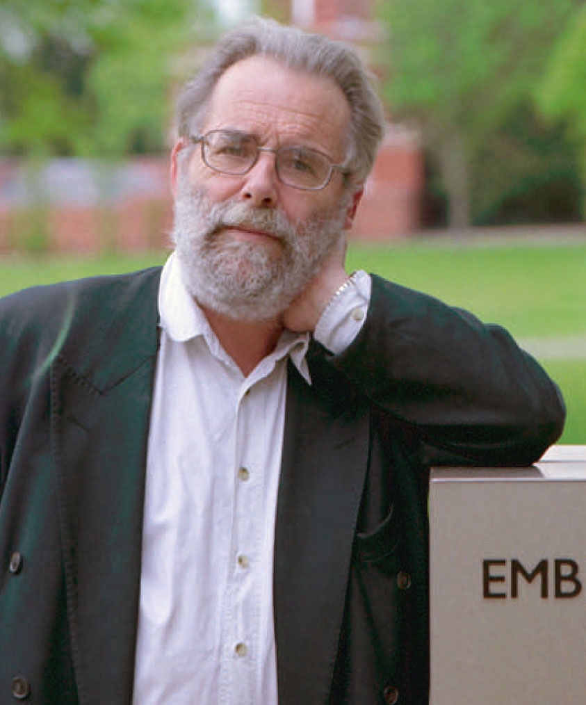 Older man with white hair, beard, and glasses, wears a white shirt and dark jacket. He rests one hand on his neck and leans against a surface with EMB visible. Blurred greenery and red-brick structure in the background.