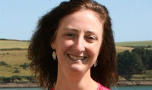 Smiling woman with curly hair wearing earrings, standing outdoors with a scenic landscape of rolling hills and trees in the background.