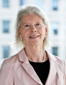 Elderly woman with short white hair, wearing a light pink blazer, black top, pearl necklace, and earrings, smiling at the camera. She is standing in front of a blurred background with large windows.