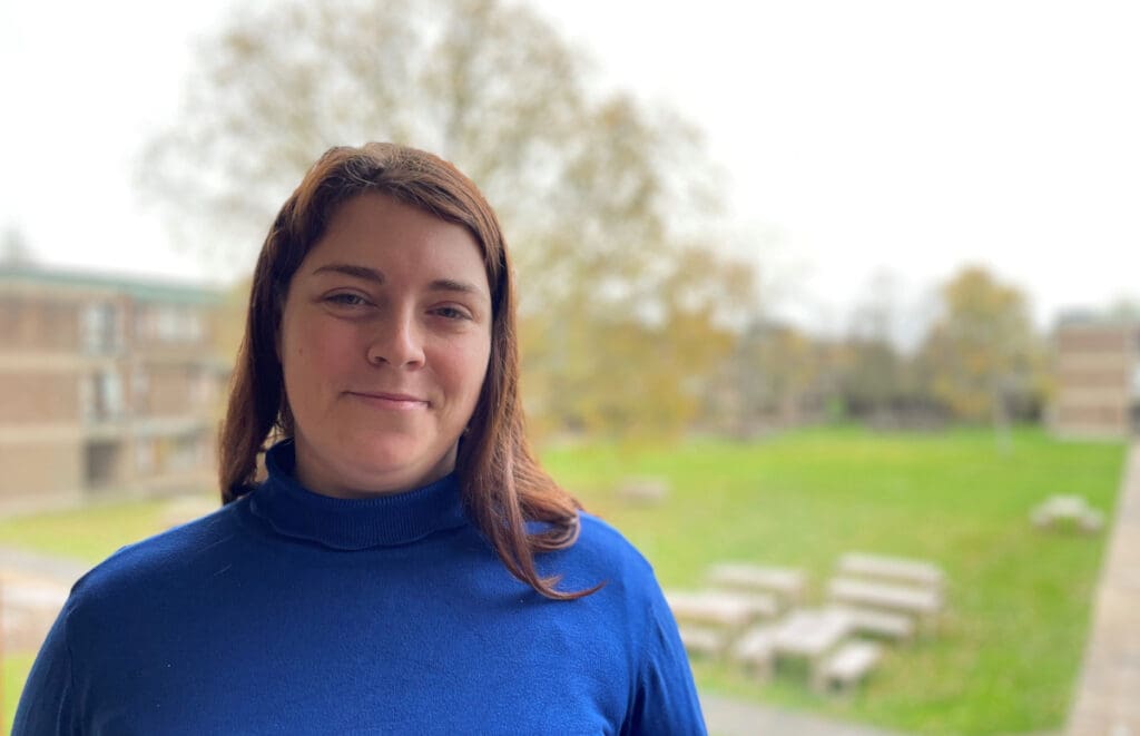 A person wearing a blue sweater is smiling while standing outside. The background features a grassy area with picnic tables and trees, with buildings on either side. The scene is bright and appears to be during the day.