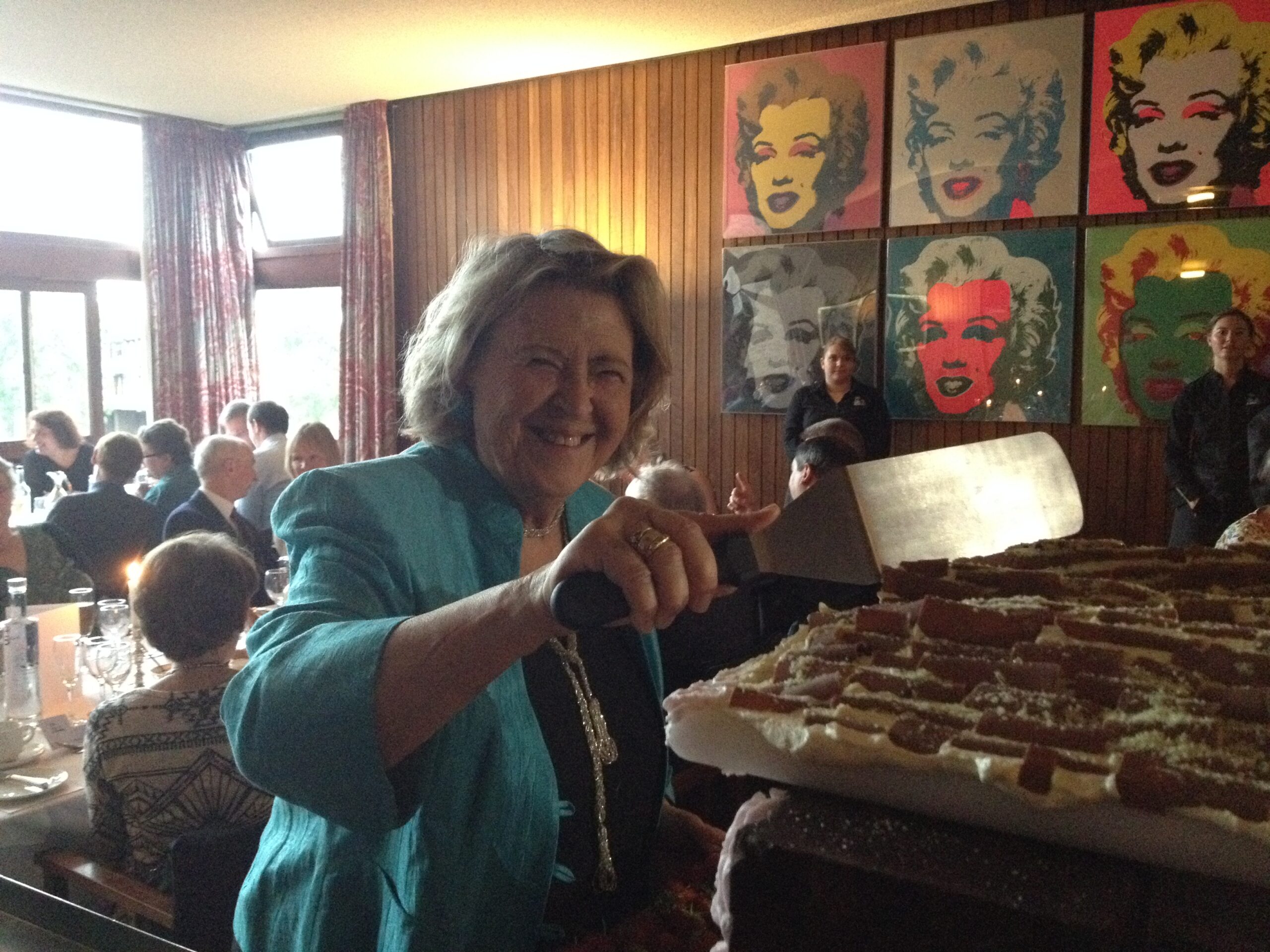Smiling woman in a turquoise jacket cuts a large cake with a spatula at a gathering. People are seated at tables in the background. The wall displays colorful pop-art portraits of a womans face.