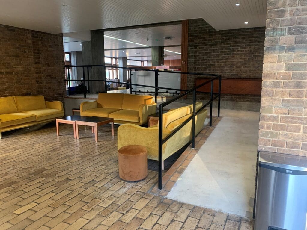Spacious room with brick walls and floor, featuring yellow sofas and a wooden table, separated by a black railing. Natural light streams in through large windows.