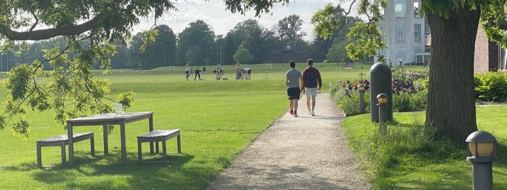 Two people walk on a path through a grassy park with trees and a wooden picnic table nearby. In the background, a group plays on the grass, and a large building and more trees are visible. The sky is partly cloudy.