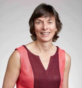 A person with short, brown hair is smiling at the camera. They are wearing a sleeveless top with red and pink panels. The background is a plain, light gray color.