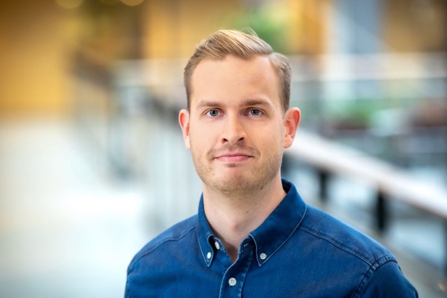 A person with short, light brown hair and a trimmed beard is wearing a blue button-up shirt. He is standing indoors with a blurred background of a building interior, giving a professional appearance.