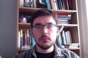 A person with short brown hair, glasses, and a beard is looking into the camera. They are standing in front of a bookshelf filled with various books. The lighting is natural, coming from a window on the right side.