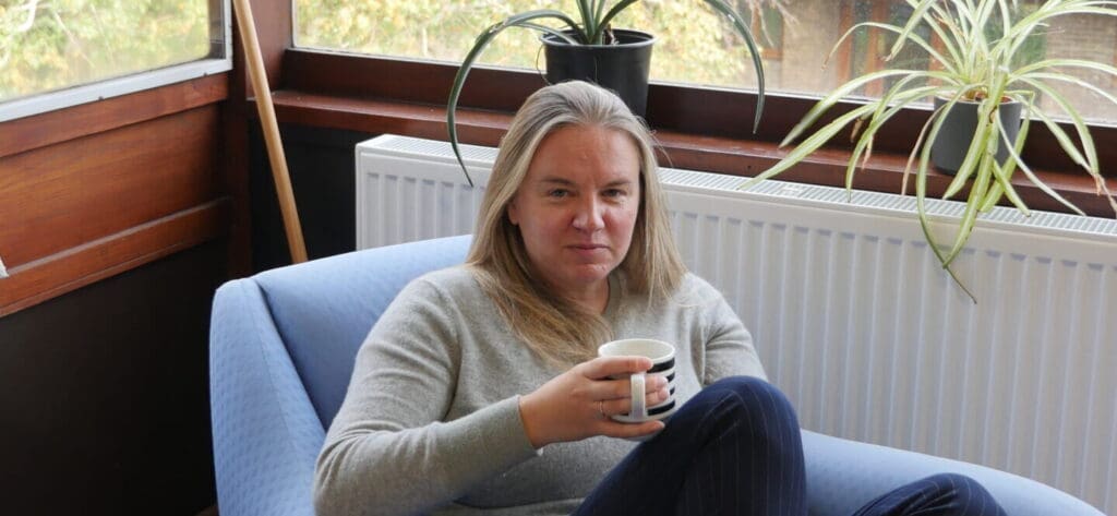 A person with long hair sits in a cozy chair holding a mug, with a relaxed expression. They are in a room with large windows and potted plants visible in the background. The setting appears calm and comfortable.
