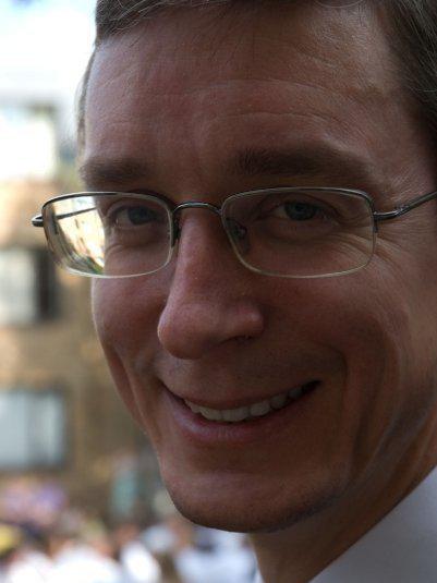 A man with glasses is smiling at the camera. He has short hair and is wearing a light-colored shirt. The background is blurred, with hints of buildings and people.