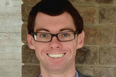 A man with short dark hair and glasses smiles at the camera, standing in front of a brick wall. They are wearing a dark suit jacket over a shirt.