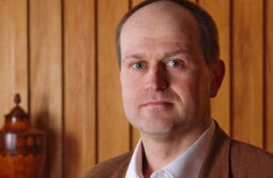 A man with short hair is wearing a white shirt and brown jacket, standing in front of a wooden paneled wall. A blurred, ornately carved wooden object is in the background.
.