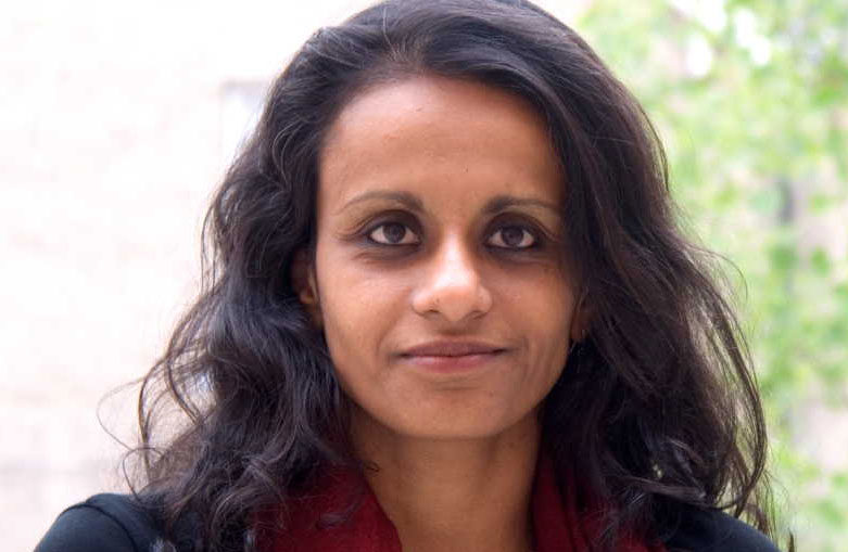 A person with long, dark wavy hair is looking at the camera with a slight smile. They are wearing a dark outfit with a maroon scarf. The background is blurred with hints of greenery.