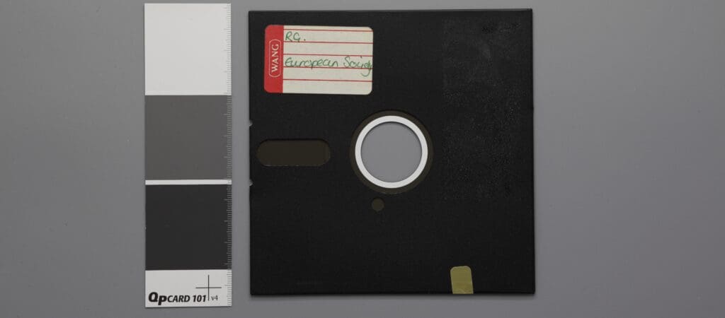 A black 8-inch floppy disk with a labeled sticker on it is shown next to a grayscale color chart. The disk features a circular metal hub. The label reads RC- and European with partial text visible.