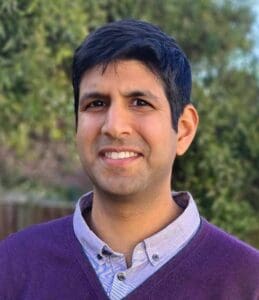 A person with short dark hair is smiling outdoors. He is wearing a purple sweater over a light-colored button-up shirt. The background features blurred greenery.