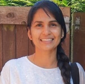 A person with long dark hair in a braid is smiling while standing in front of a wooden fence and green foliage, wearing a white shirt and carrying a black bag over their shoulder.