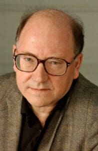 A man with glasses and a balding head is wearing a brown blazer and a dark shirt and looking directly at the camera with a neutral expression. The background is plain and light-colored.