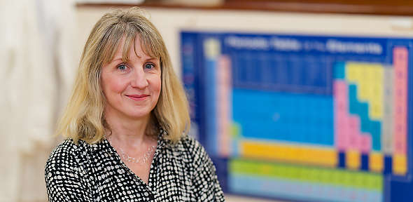 A woman with blonde hair and a patterned blouse smiles at the camera. Behind her, there is a blurred periodic table poster.