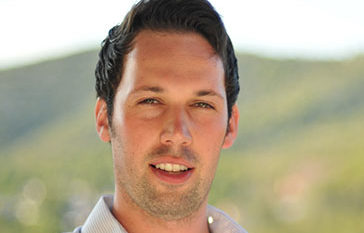 A man with short dark hair is smiling slightly. He is wearing a light-colored collared shirt. The background is a blurred, natural landscape with hills and greenery.