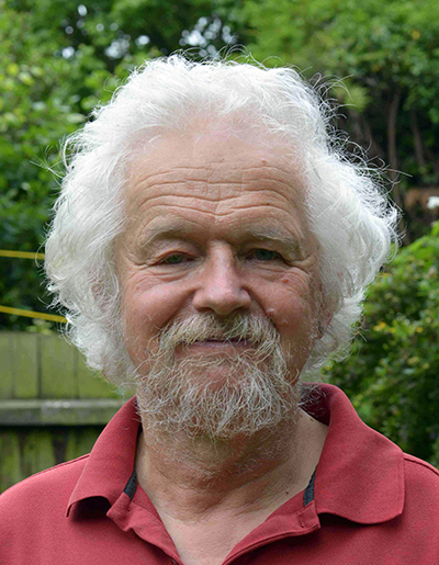 An man with white hair and a beard is smiling at the camera. He is wearing a red polo shirt. The background features green foliage and a wooden fence, suggesting an outdoor setting.