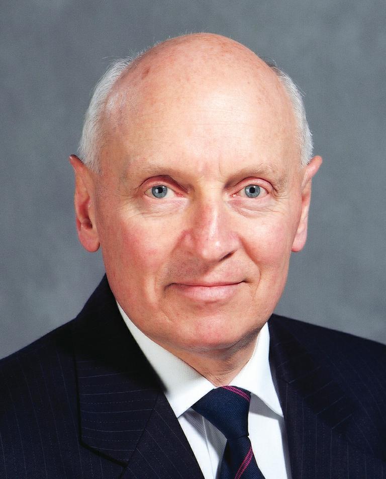 A balding man in a dark pinstripe suit and tie, with light skin, is looking directly at the camera against a gray background.