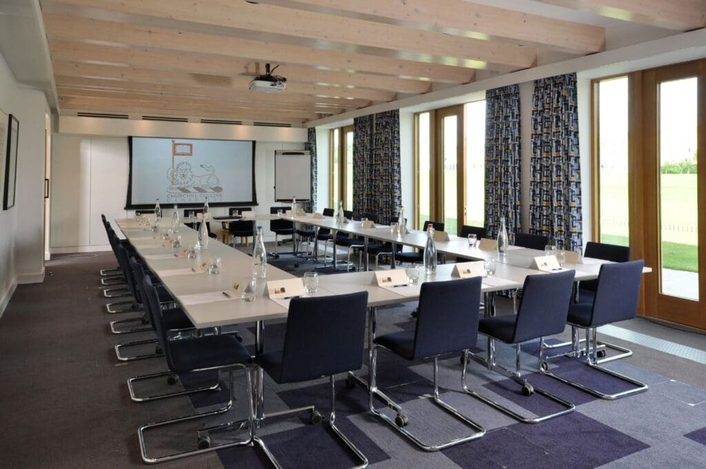 A modern conference room with U-shaped setup, featuring tables with water bottles and name cards, surrounded by black chairs. A projector screen and flipchart stand at the front. Large windows on the right provide natural light.