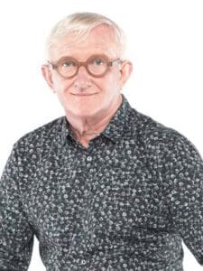 Man with short white hair and round glasses, wearing a patterned dark shirt with a subtle floral design, smiles at the camera against a white background.