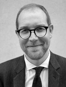 A man with short hair, wearing black-rimmed glasses, a suit, and a tie, smiles at the camera against a plain background. The image is in black and white.