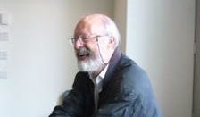 A man with a beard and glasses is smiling and looking to the right. He is wearing a dark jacket over a white shirt. The background is a pale wall with some indiscernible text.