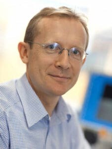 Man wearing glasses and a checkered shirt, smiling slightly. He is in an office setting with a blurred blue screen in the background.