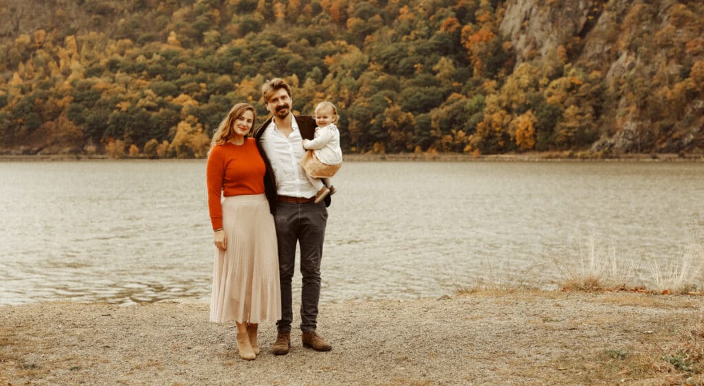 A family stands by a lake with a forested hillside in the background. The woman wears a red sweater and beige skirt, the man is in a dark jacket and holds a child in a light outfit. The leaves on the trees are in autumn colors.