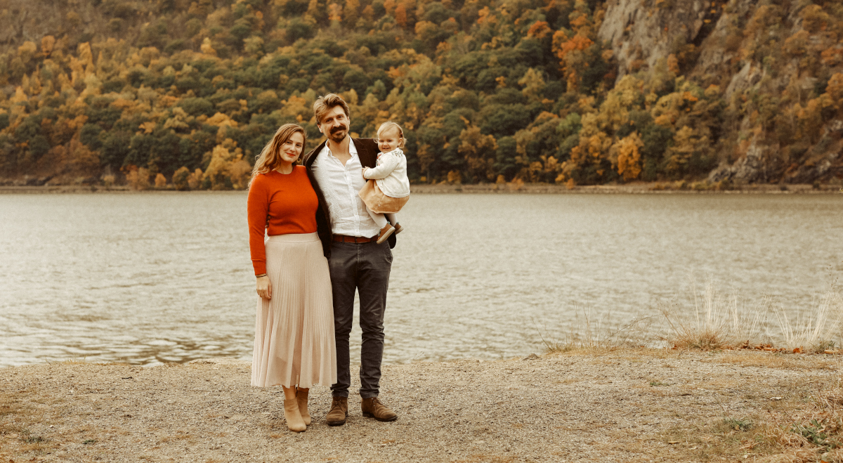A family stands by a lake with a forested hillside in the background. The woman wears a red sweater and beige skirt, the man is in a dark jacket and holds a child in a light outfit. The leaves on the trees are in autumn colors.