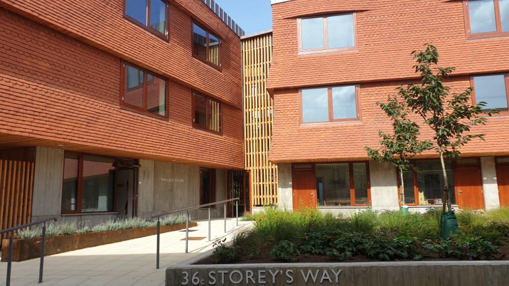 Exterior view of a modern building with red brick and wood paneling. The entrance sign reads 36 Storeys Way. There are windows, a small tree, and planters in the courtyard, set against a partly cloudy blue sky.