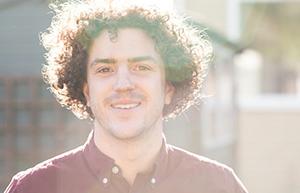 Smiling man with curly hair wearing a maroon shirt, standing outside with sunlight creating a warm, glowing effect in the background.