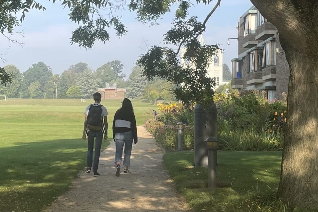 Two people walk along a tree-lined path beside a building with colorful flowers. The sky is mostly clear, and green fields stretch out in the background, suggesting a calm, sunny day.