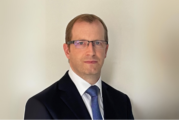 A man with short hair is wearing glasses, a black suit, a white shirt, and a blue tie. He is standing against a plain light-colored background, looking directly at the camera with a neutral expression.