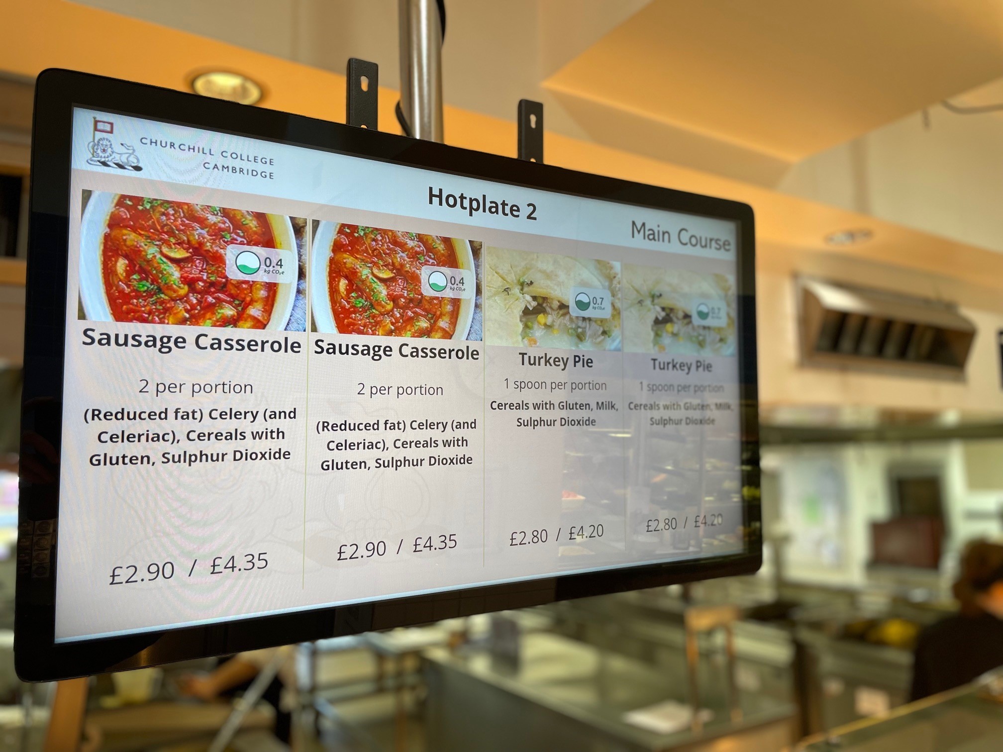 A digital menu at Churchill College, Cambridge, displays two main courses: Sausage Casserole and Turkey Pie, with nutritional information and prices. The background shows a food service area with a person in uniform.