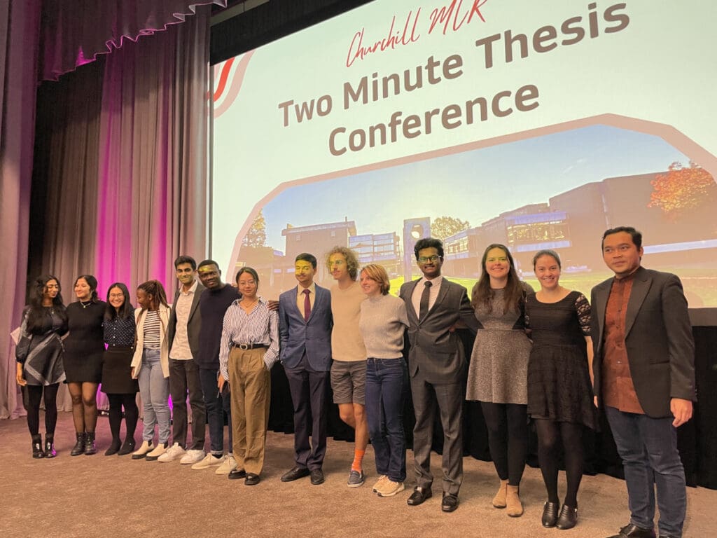 A group of people stand on stage in front of a screen that reads Churchill MCR Two Minute Thesis Conference. They are posing for a photo, dressed in formal and casual attire. The background features a large building image.