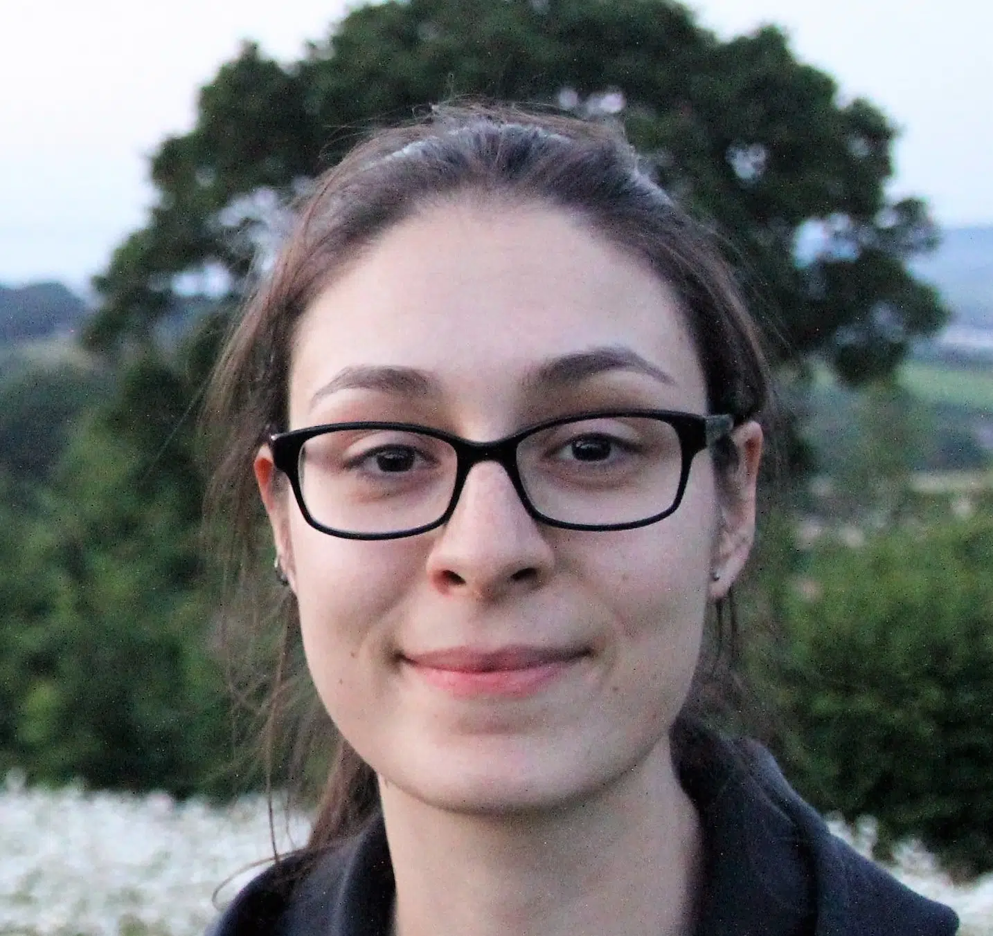 A person with long brown hair and black glasses is smiling slightly. A blurred background features a large tree and greenery. The sky appears to be overcast or near dusk.