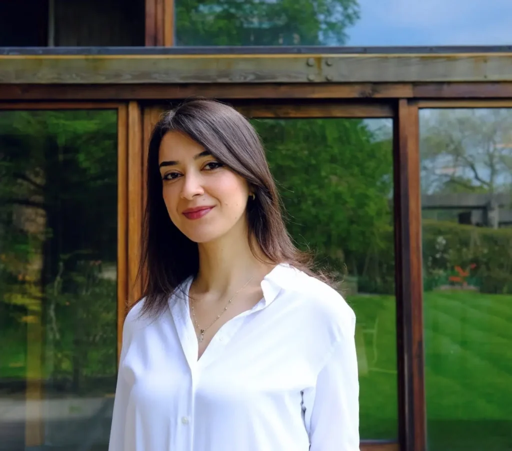 Woman with long brown hair and a white blouse smiling in front of a large window with a view of a lush green garden in the background.