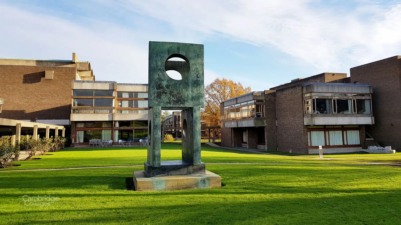 A modern abstract sculpture stands on a green lawn surrounded by contemporary brick buildings. The sculpture features geometric shapes and a circular opening. The sky is partly cloudy, and a tree with autumn leaves is visible in the background.