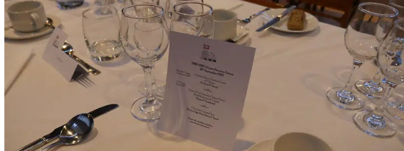 A neatly set dining table with multiple wine and water glasses, a menu card, cutlery, and a folded napkin. Theres also a tea cup and saucer, with a small pastry on a separate plate in the background.