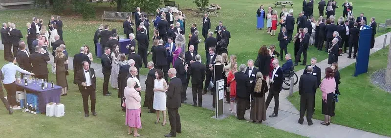 A large outdoor gathering of people dressed in formal attire, mingling on a grassy area. Some are standing in groups while others are near a table with drinks. Trees and benches are visible in the background.