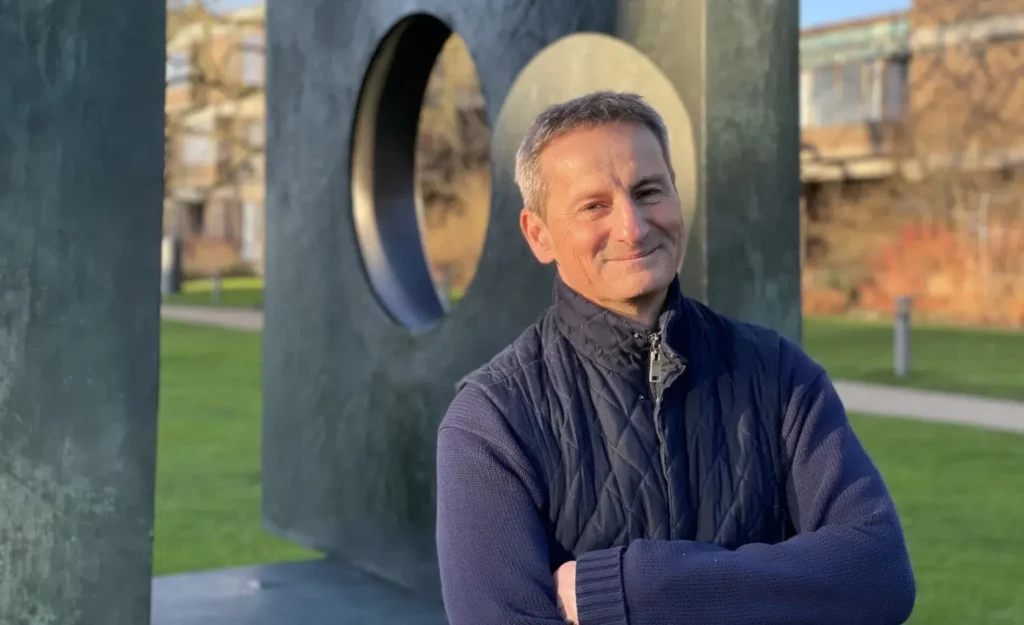 A person stands outdoors, smiling with arms crossed, wearing a dark quilted jacket and blue sweater. Behind him is a large modern sculpture with circular cutouts, set against a grassy area and building in the background.