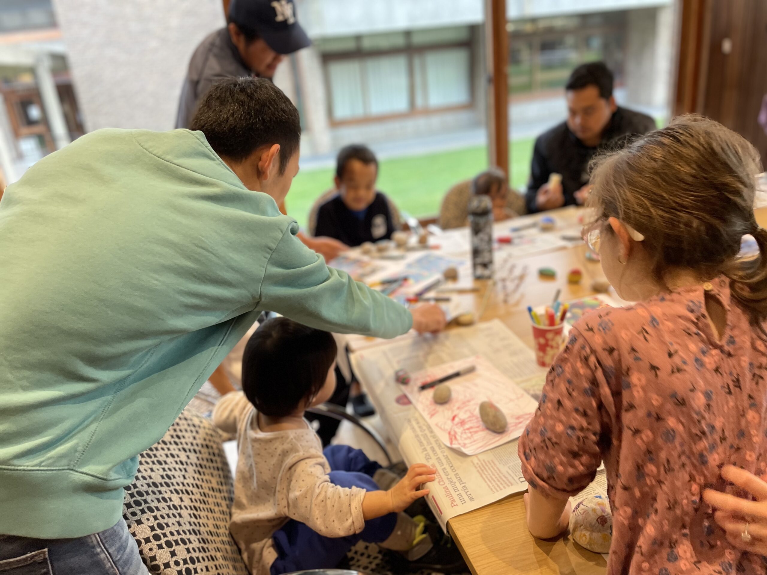 A group of children and adults sit around a table indoors, painting and drawing on rocks and paper. Art supplies, colored markers, and brushes are scattered on the table. The scene is lively and collaborative.