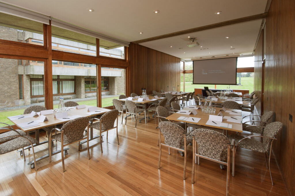 A modern conference room with large windows, wooden floors and walls, several tables and chairs arranged for a meeting, and a projector screen displaying a presentation at the front.