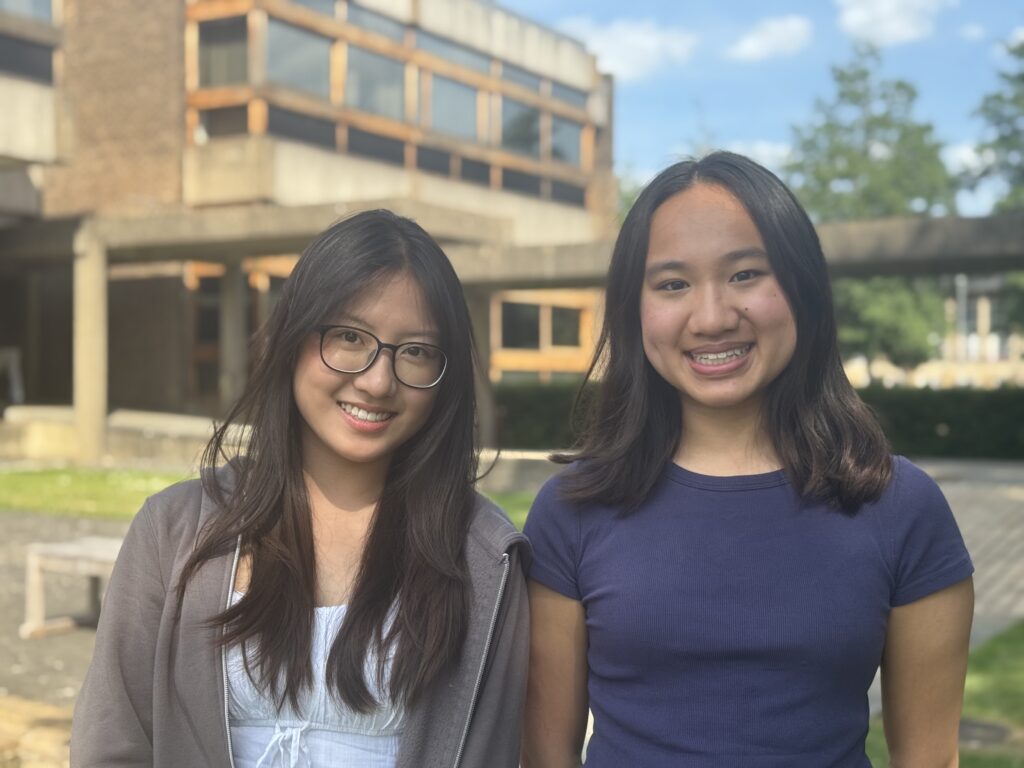 Two young women with dark hair are standing outside a modern building smiling at the camera