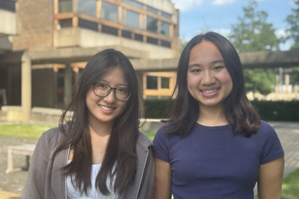 Two young women with dark hair are standing outside a modern building smiling at the camera