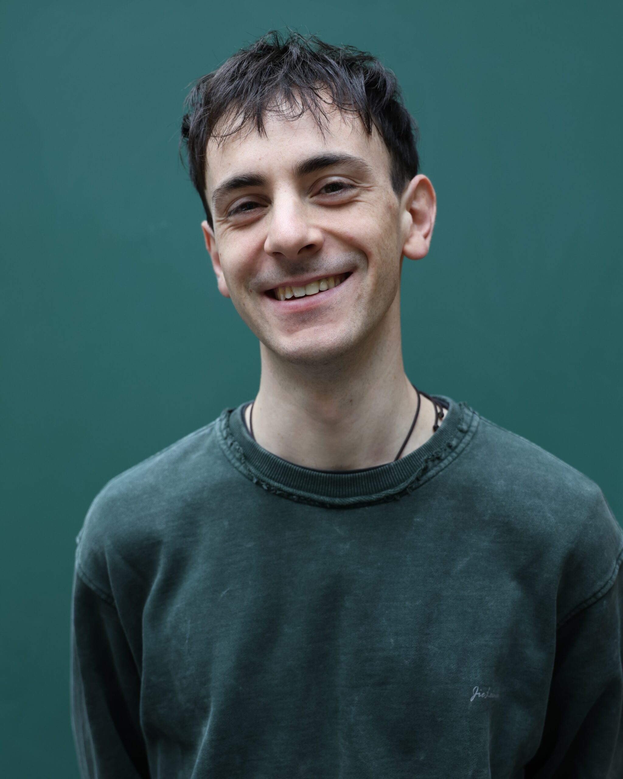 A young man with short dark hair, wearing a faded green sweatshirt, smiles while standing in front of a plain green background.